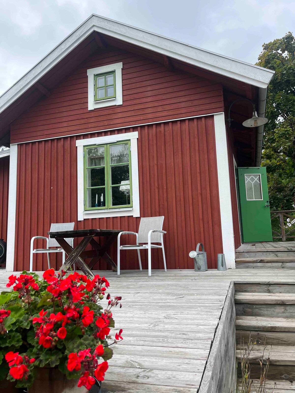 A red wooden cottage features a green door and a large window. A small table with two chairs is positioned on a wooden deck. Bright red flowers in a planter add a pop of color to the entrance, welcoming visitors.