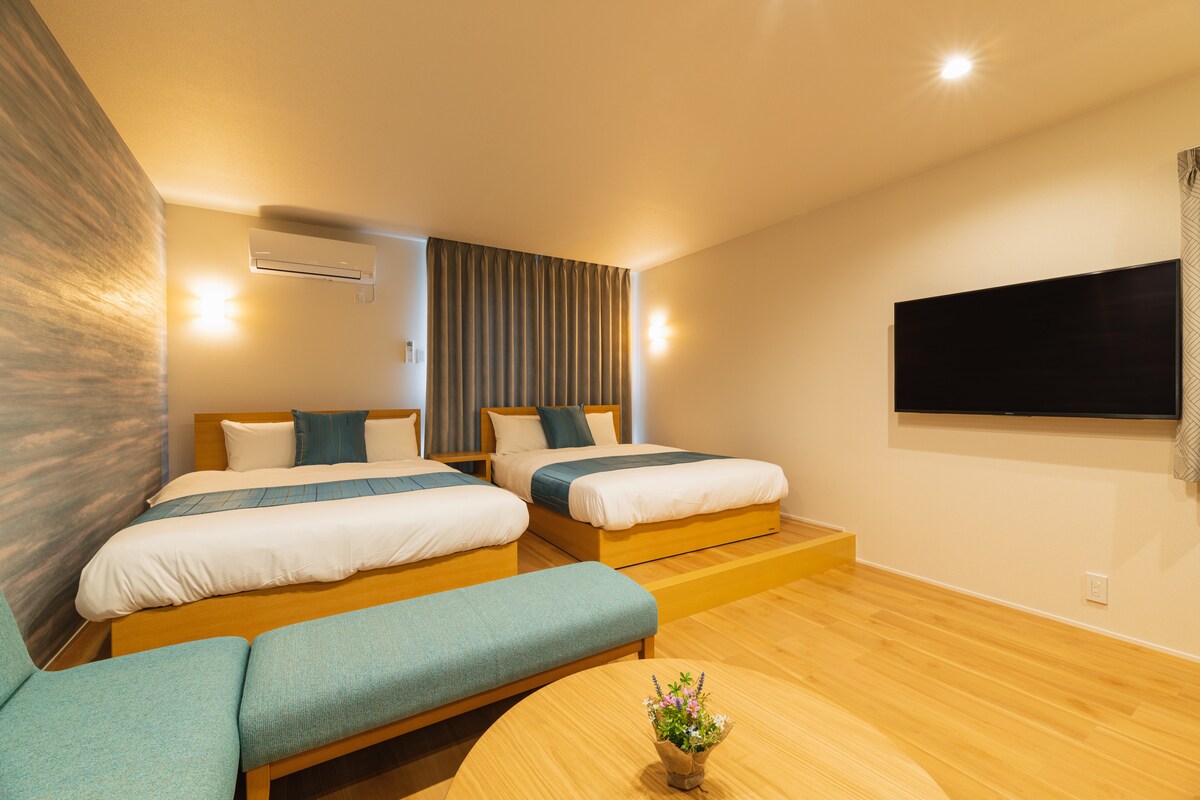 A spacious room features two double beds with light blue accents. Soft natural light enters through the curtains, illuminating the minimalist decor. A small floral arrangement sits on the round wooden table, while a television is mounted on the wall opposite the beds.