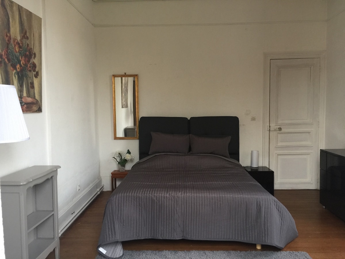 A spacious bedroom features a large, neatly made bed with a textured gray blanket, complemented by neutral-toned walls. A mirror reflects natural light, while a small table and lamp sit beside the bed. A modern cabinet is positioned against the opposite wall.