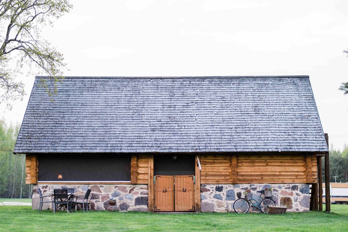 A rustic summer house is presented, featuring wooden and stone construction. The seamless roof is gently sloped, and an inviting porch includes seating for relaxation. A bicycle is parked beside the house, surrounded by lush green grass and open space, enhancing the peaceful rural setting.
