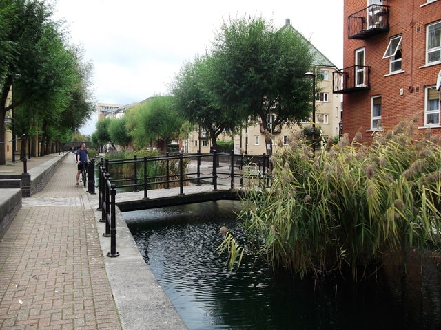 A serene canal scene features a pedestrian bridge spanning over calm waters, surrounded by lush greenery. On either side, pathways are lined with trees, and residential buildings are visible in the background, creating a tranquil urban environment.