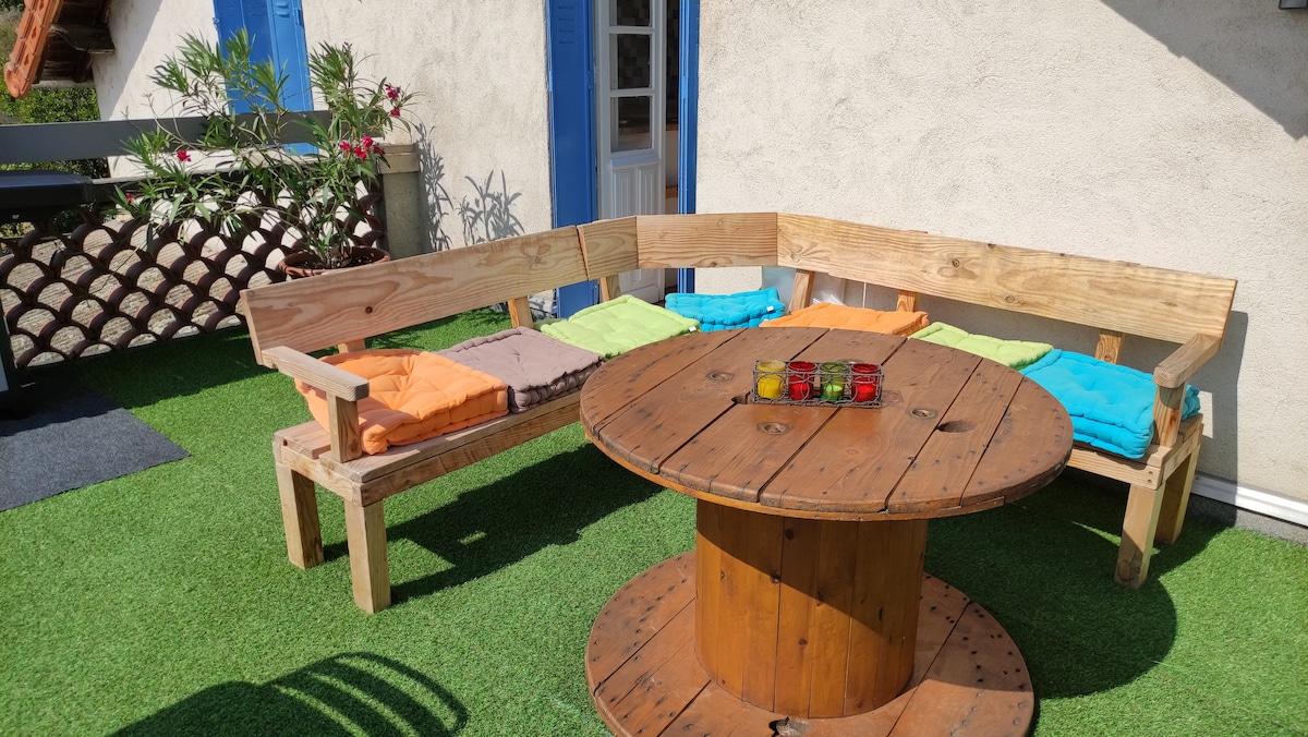 An outdoor seating area features a circular wooden table surrounded by a rustic wooden bench. Colorful cushions in shades of orange and teal enhance the seating. Green artificial grass covers the ground, complemented by potted plants in the background.