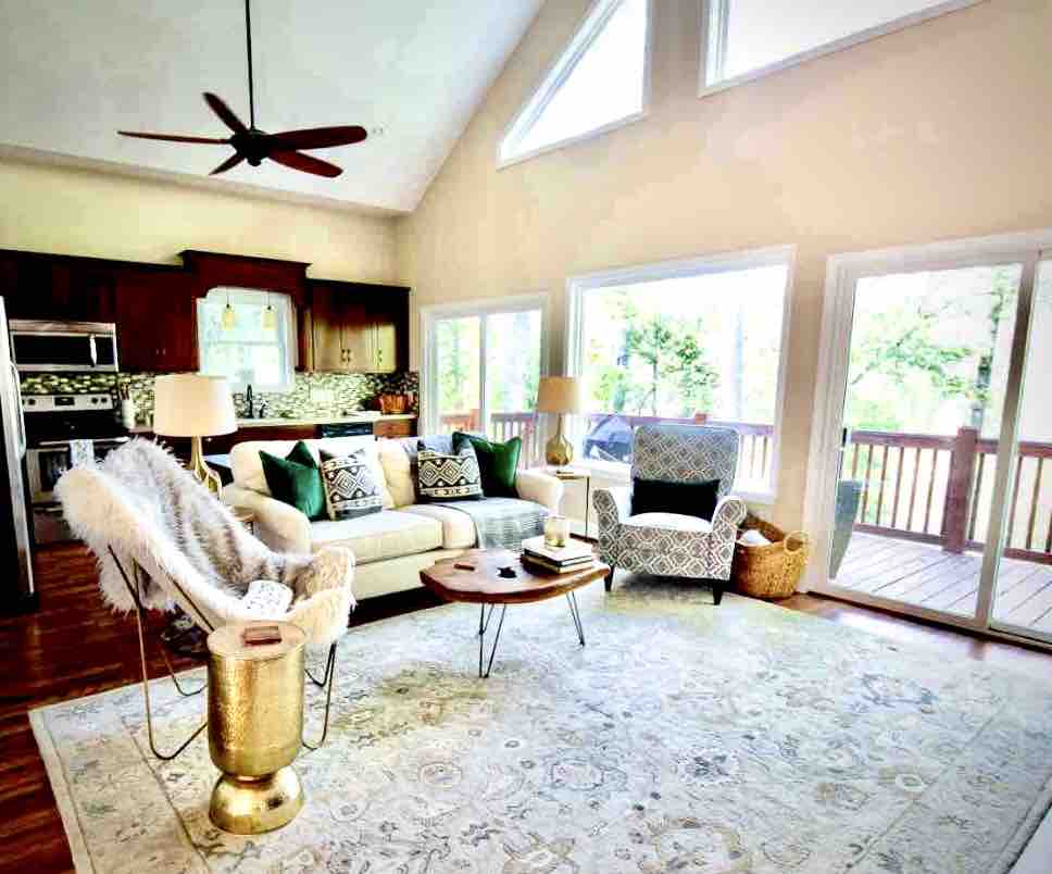 A comfortable living area features large windows allowing natural light to fill the space. A neutral sofa with decorative pillows is complemented by a stylish accent chair and a central coffee table. A modern ceiling fan is mounted above, enhancing the open, airy atmosphere.