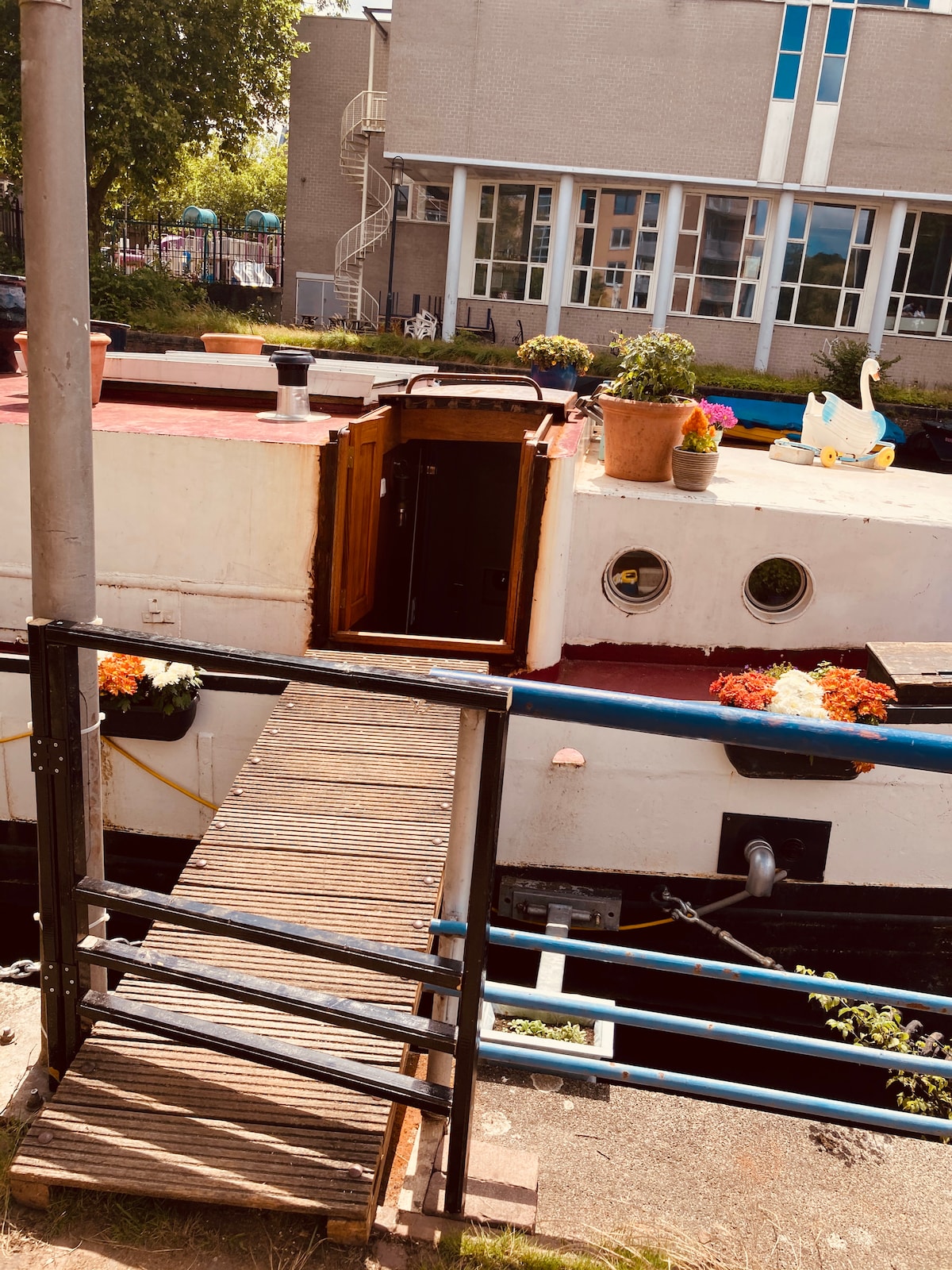 The entrance to the houseboat is depicted, featuring a wooden ramp leading to a door surrounded by potted plants. The vibrant flowers add color to the exterior, while the boat's structure is complemented by windows and a spiral staircase seen in the background.