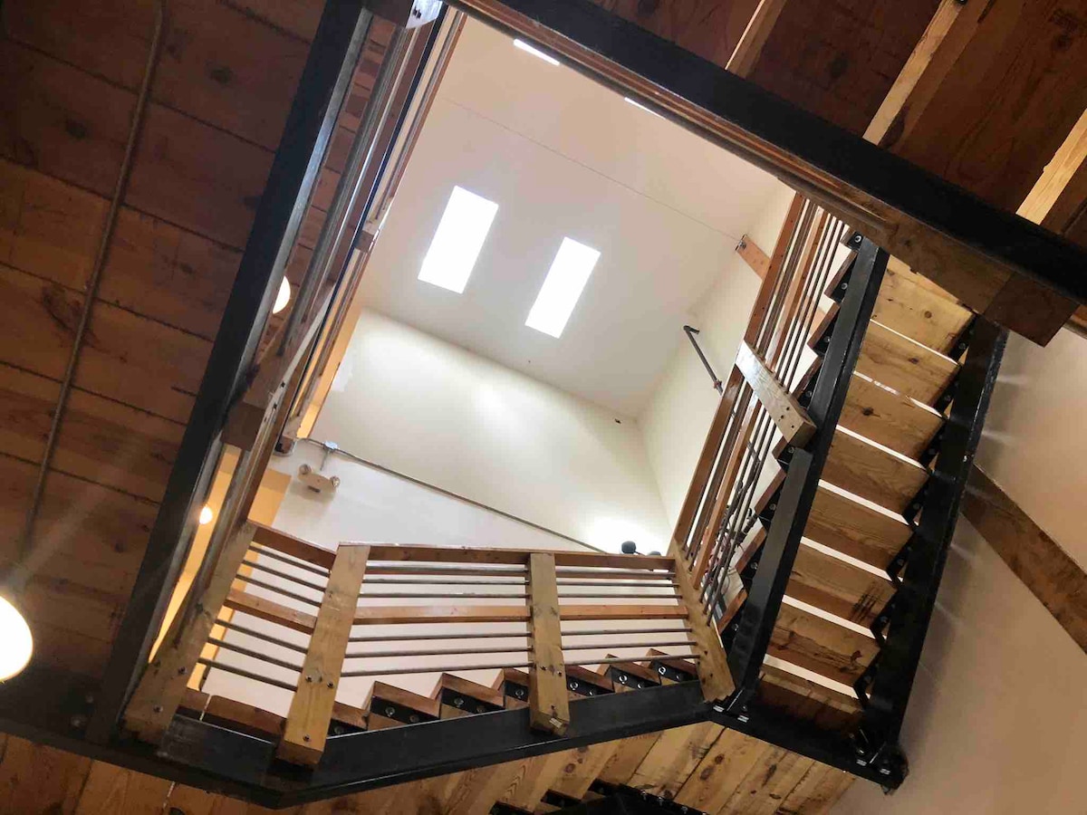 A view from the lower level captures a wooden staircase transitioning to the upper section, featuring metal railings and a spacious landing. Skylights create an airy atmosphere, allowing natural light to enhance the warm tones of the exposed wooden beams.