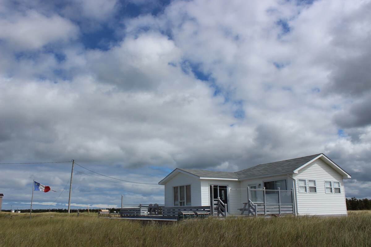 The beach cabin is shown situated among natural dune grass, with a large deck extending from the exterior. Soft gray clouds drift across the sky, complementing the white exterior of the cabin. A flag is visible nearby, gently waving in the breeze.