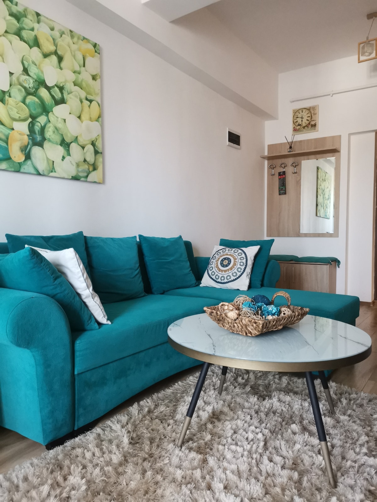 A bright living area is presented with a turquoise sofa adorned with cushions, complemented by a round coffee table. A decorative bowl filled with colorful items rests on the table. A large artwork featuring vibrant green tones decorates the wall, enhancing the cheerful ambiance.