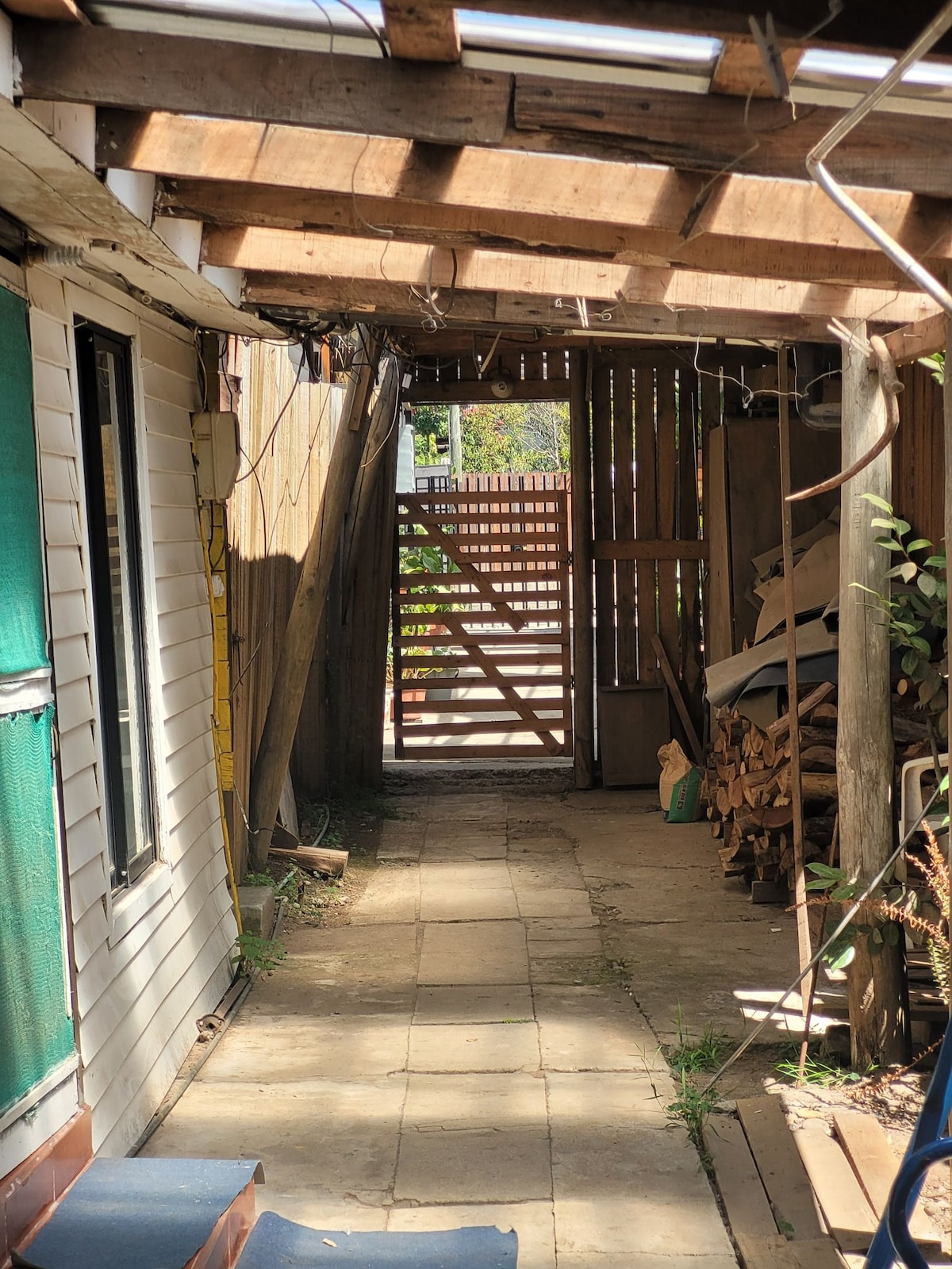 A narrow outdoor corridor is visible, lined with wooden beams overhead. The path is paved with square tiles and extends towards an open gate at the end. On one side, stacked wood and some greenery are present, while the opposite side features a white-walled structure with a green door.