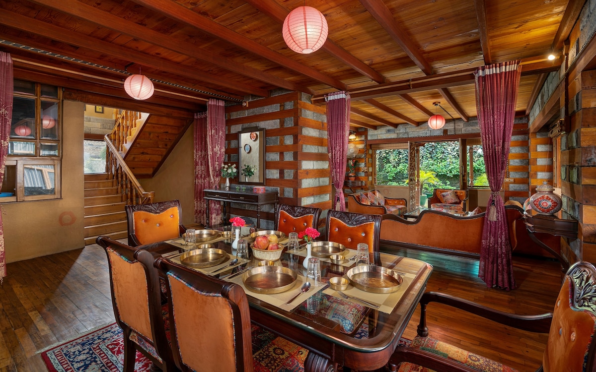 An inviting dining area features a polished wooden table set with metal plates and bowls, complemented by plush orange chairs. Large windows allow natural light to fill the space, revealing glimpses of the vibrant garden outside. Hanging lanterns add a warm touch to the wooden-beamed ceiling.