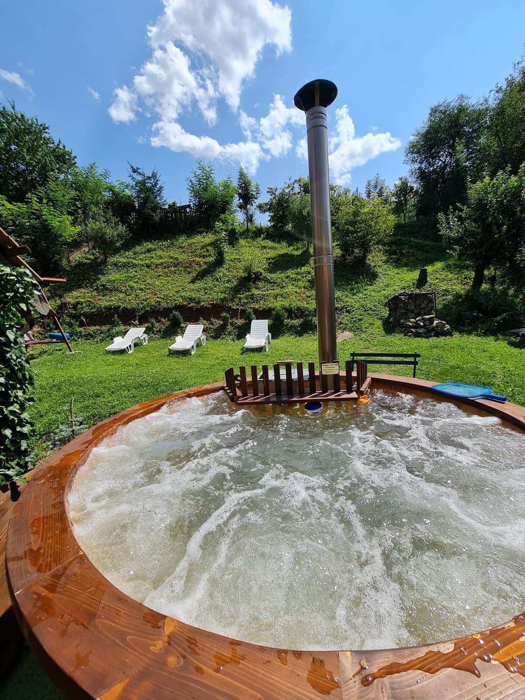 A round wooden hot tub is seen, bubbling gently under a clear blue sky. Behind the hot tub, a grassy area features several white lounge chairs for relaxation. Distant trees provide a natural backdrop, enhancing the outdoor space.