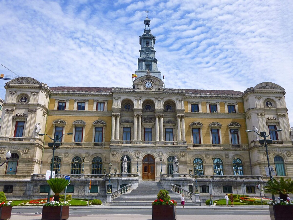The historic building features an ornate façade with a grand staircase leading to the entrance. Statues and detailed architectural elements adorn the exterior. A clocktower provides an eye-catching focal point, while clouds drift across the sky above, adding depth to the scene.