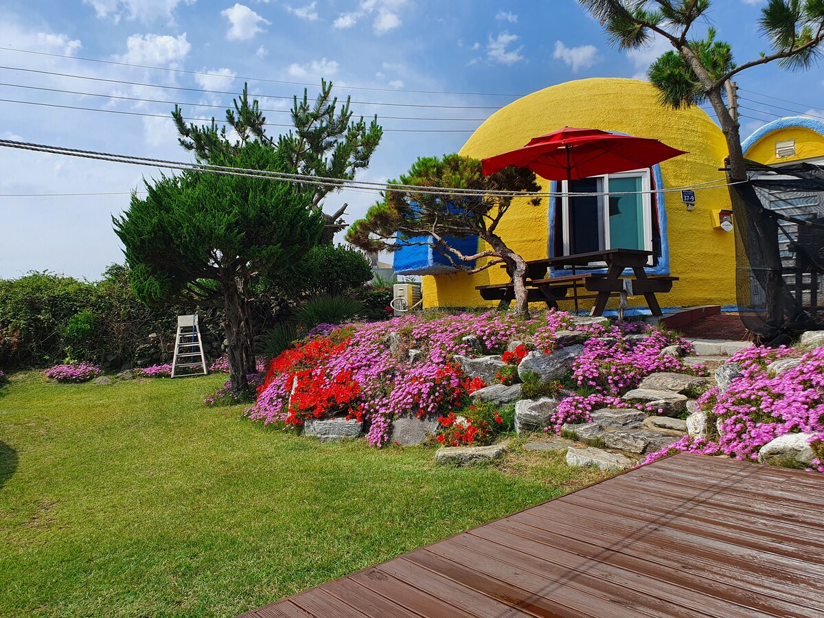 A vibrant yellow structure is framed by a colorful garden filled with various flowers. A wooden deck leads to the entrance, while a picnic table under a red umbrella offers an outdoor dining space. Lush greenery and carefully arranged stones enhance the natural setting.