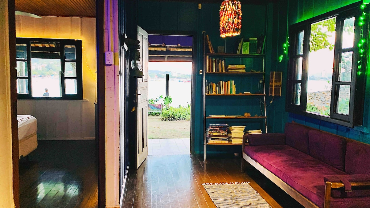 An open interior showcases a cozy living space featuring a purple sofa and a bookshelf filled with books. Natural light floods in through black-framed windows, revealing a view of the garden and the water beyond. The walls are painted in a calming blue hue.