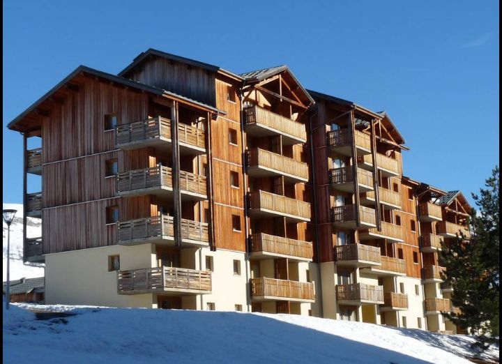 The apartment building features a contemporary design with wooden balconies and a light-colored base. Set against a clear blue sky, the structure is surrounded by snow, creating a distinct contrast with the brown wooden facades. Multiple levels are visible, enhancing its spacious appearance.