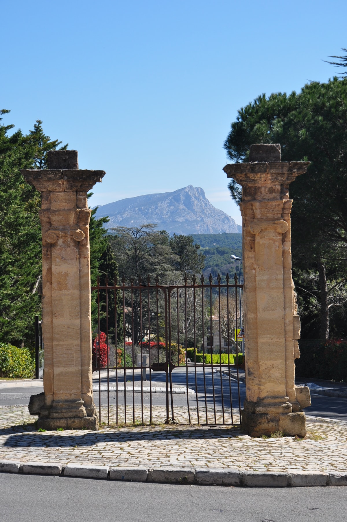 An entrance gate framed by stone columns stands prominently on a clear day. In the background, the imposing Montagne Sainte Victoire rises against a blue sky, surrounded by lush greenery and trees, creating a sense of peace and natural beauty.