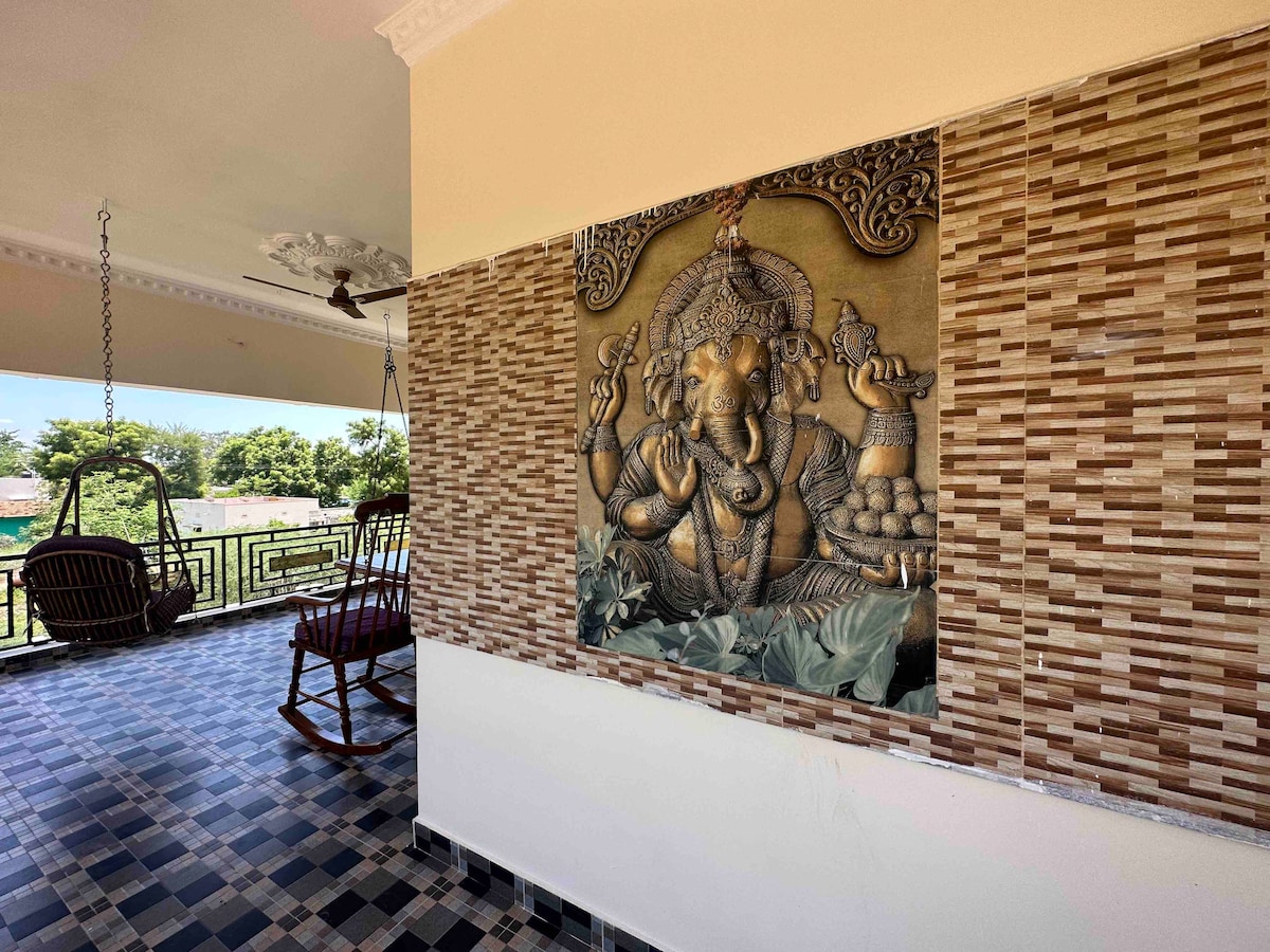 An intricately carved wall mural of Lord Ganesha is displayed, surrounded by decorative tiles. A comfortable hanging swing is positioned on the balcony, with a scenic view visible through the railing. Natural light illuminates the space, enhancing the calm ambiance.