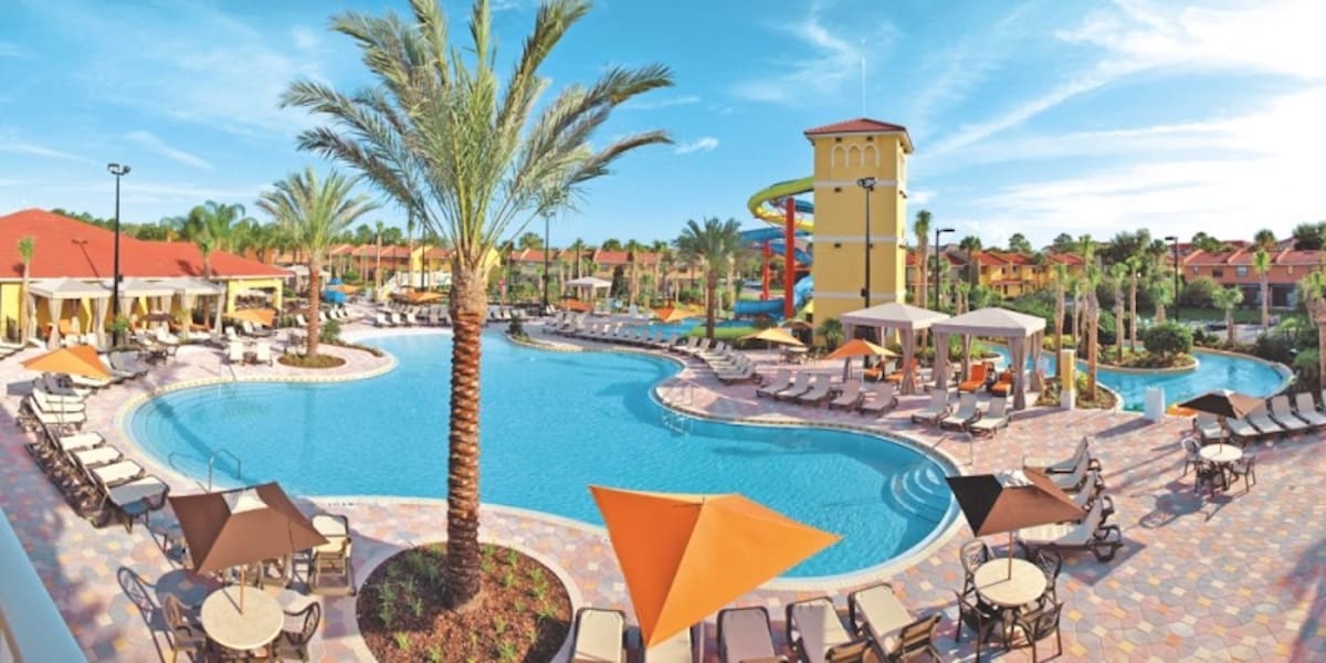 The expansive resort pool area is surrounded by palm trees and features a winding pool with lounge chairs scattered along the deck. A water slide and colorful tower are visible in the background, providing a vibrant atmosphere for relaxation and recreation.