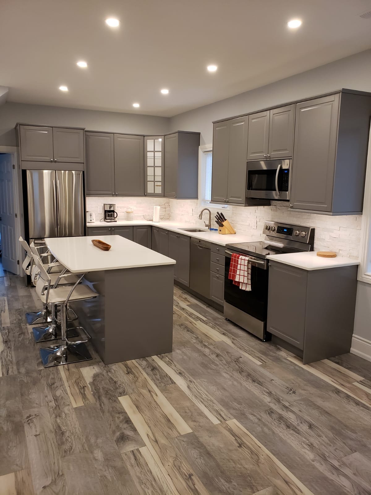 The modern kitchen features sleek gray cabinetry, stainless steel appliances, and a large island with seating for four. Soft lighting illuminates the white countertops and backsplash, while a coffee maker and other essentials are visible, providing a functional and inviting culinary space.