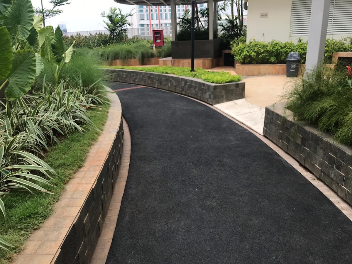 A winding pathway is framed by lush greenery and low-lying plants. The textured surface of the path is bordered by neatly arranged stone walls and a manicured garden, providing a serene setting for walking or jogging.