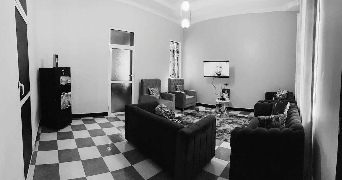 A spacious living area features a mix of comfortable seating arrangements, including two sofas and several armchairs. A large television is mounted on the wall, while natural light filters through the windows. A patterned rug adds texture to the tiled floor.