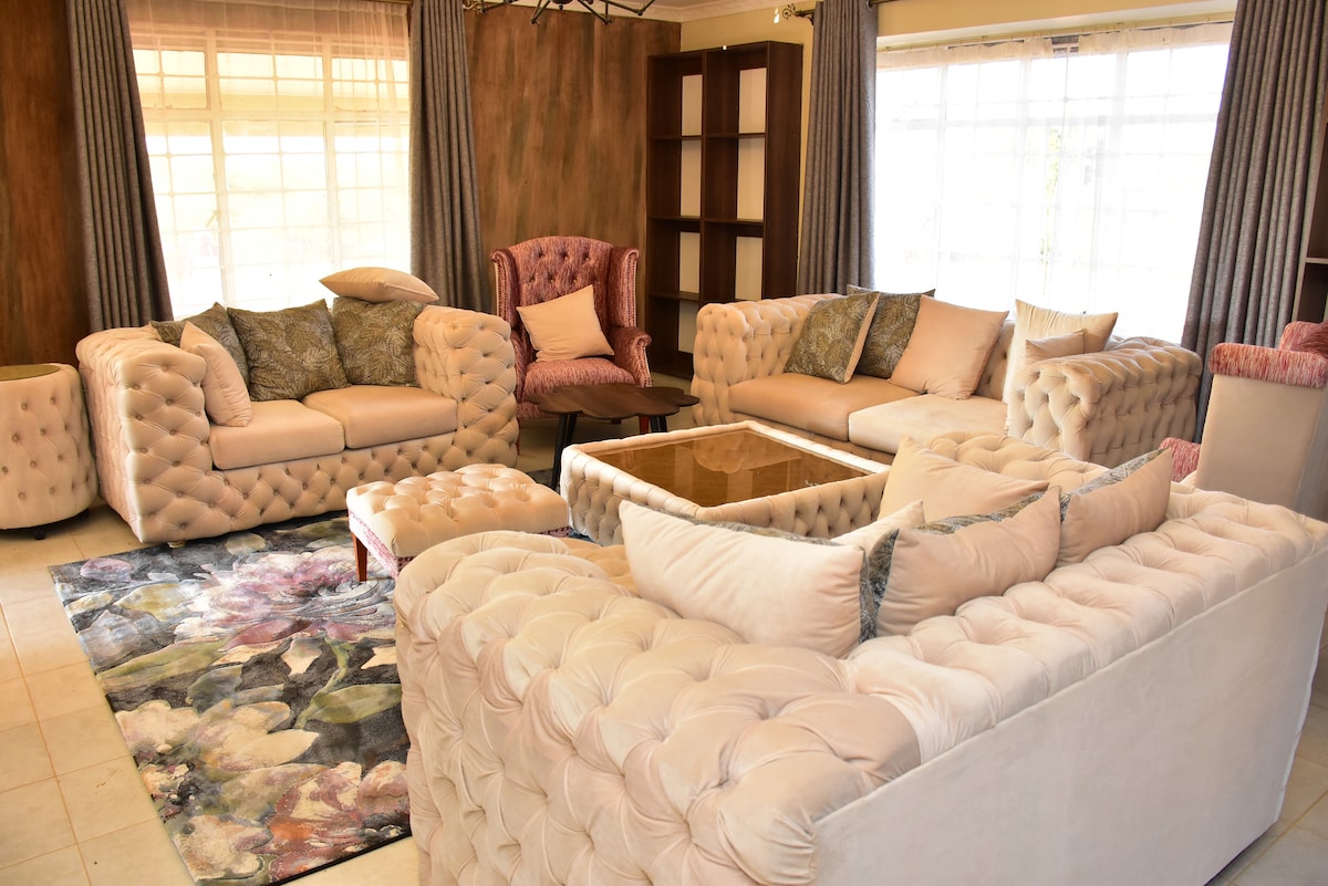 The living area features elegant tufted sofas in soft tones, arranged around a glass-top coffee table. A colorful area rug enhances the space, while large windows allow natural light to fill the room. Wing chairs provide additional seating, complemented by decorative pillows.