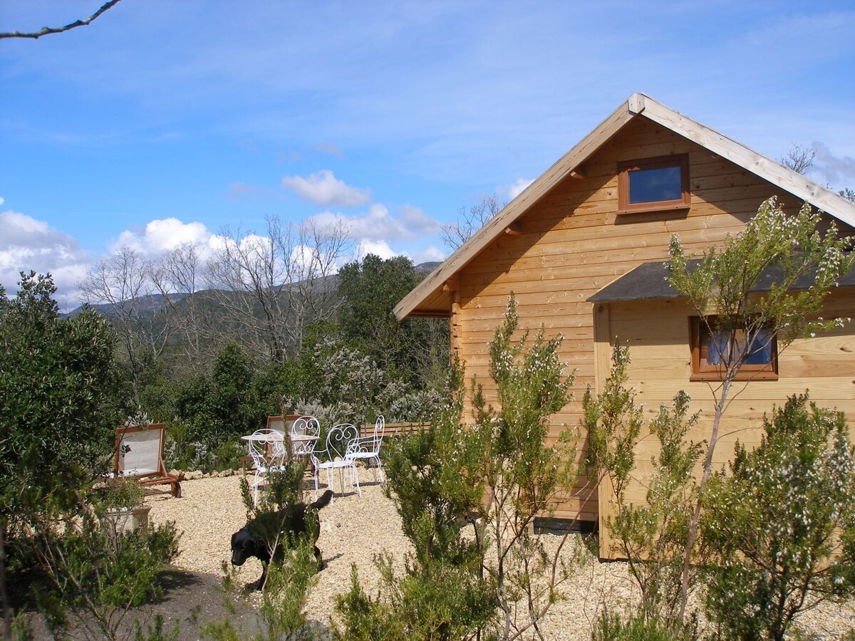 A wooden house is situated amidst natural surroundings, with a pathway leading to an outdoor seating area. The property features a view of distant mountains, framed by trees and shrubs, creating a serene and private environment.