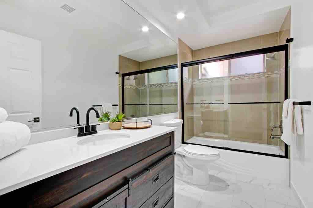A modern bathroom features a spacious vanity with a dark wood finish and a white countertop. A large glass shower enclosure is visible, along with a toilet and bright lighting. A small plant and towels are neatly arranged on the counter, enhancing the fresh atmosphere.