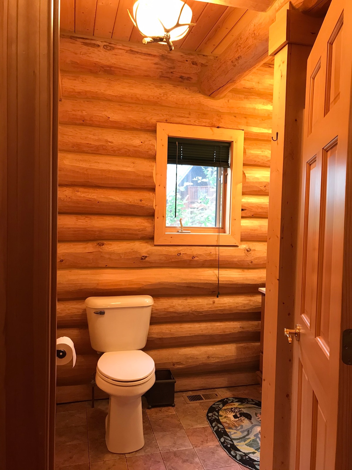 The private bathroom features natural wood paneling and a small window allowing light to enter. A white toilet is positioned against the log wall, and a decorative rug is placed on the tile floor near the toilet. A doorway leads to the rest of the suite.