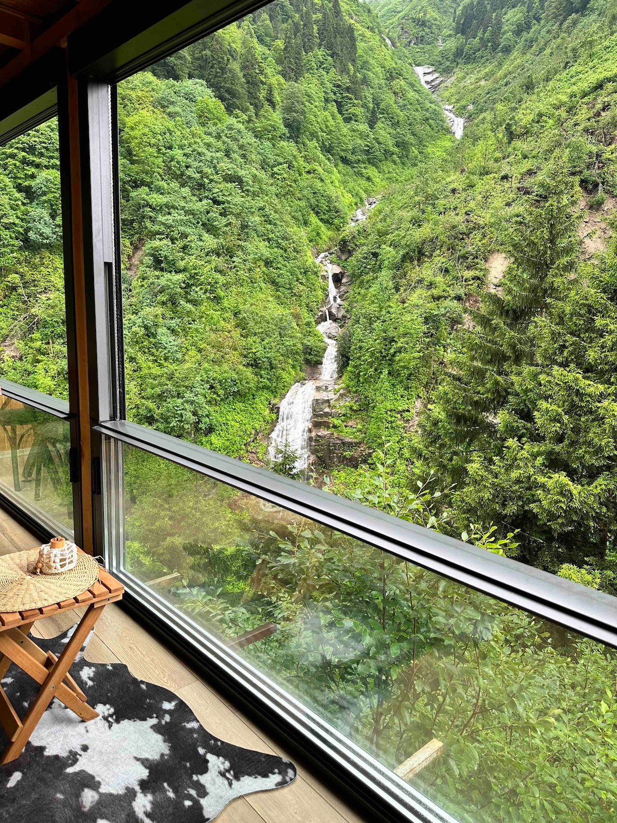 A large window frames a stunning view of a lush green landscape with cascading waterfalls. A comfortable chair and a patterned rug are positioned nearby, inviting guests to relax and enjoy the natural beauty outside.