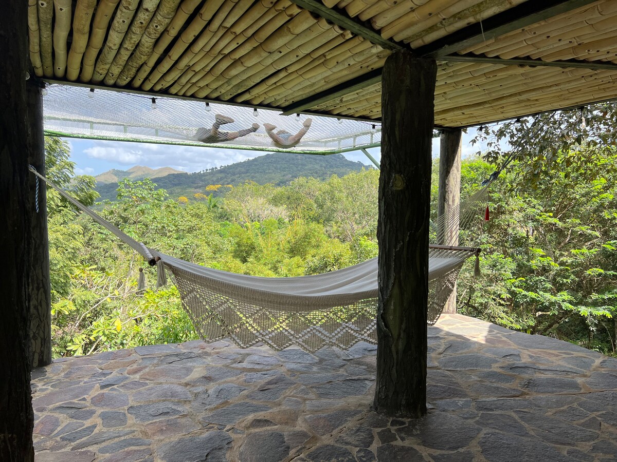 A hammock is suspended between wooden posts on a stone floor, offering a peaceful spot to relax. Lush greenery surrounds the area, with a view of distant mountains visible through the trees. Natural light filters through, enhancing the tranquil setting.