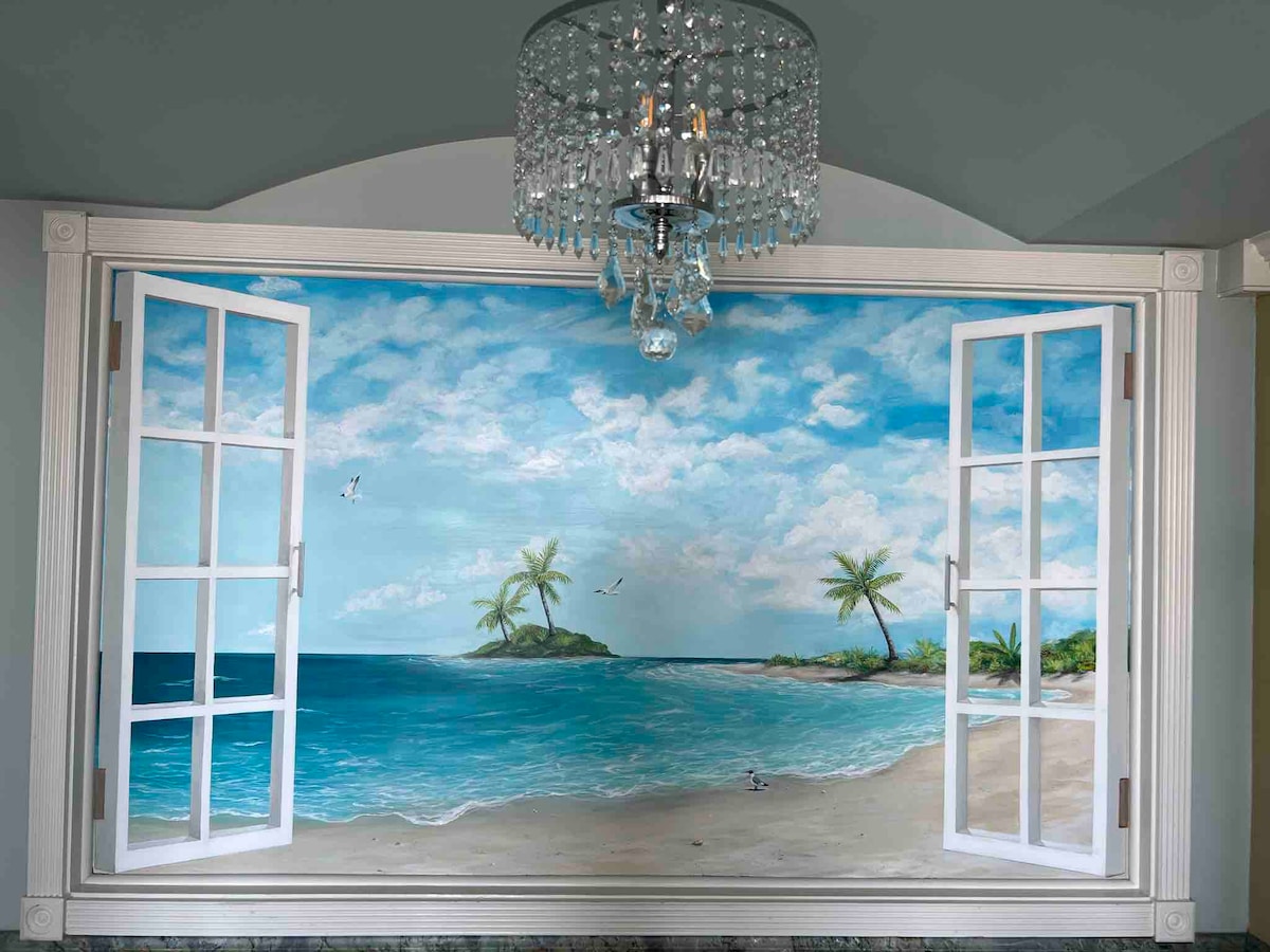 A mural depicting a serene beach scene is framed by white open window shutters. The artwork features a calm ocean, sandy shore, and palm trees under a blue sky with fluffy clouds. A crystal chandelier hangs above, adding a touch of elegance to the space.