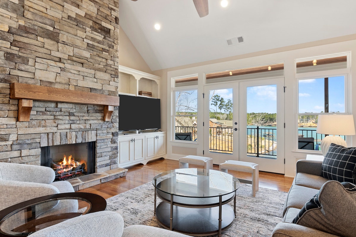 A spacious living area features a stone fireplace complemented by a wooden mantel. Large glass doors provide access to the balcony, revealing scenic views. Comfortable seating arrangements surround a circular coffee table, with natural light filling the room through large windows.