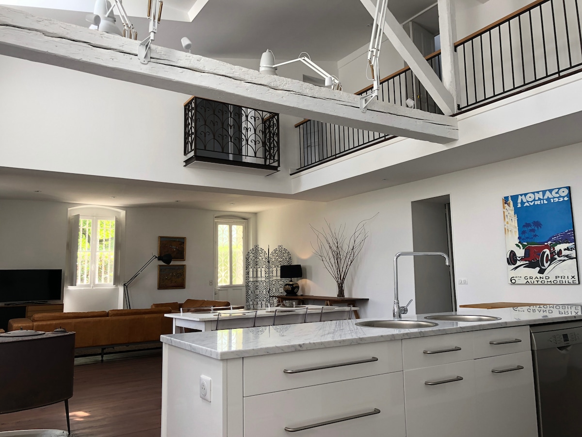 A modern open-concept living space features a sleek kitchen island with a double sink. Large windows allow natural light to fill the room, highlighting a comfortable seating area with a brown sofa and a dining table set against a backdrop of white walls and elegant decor.