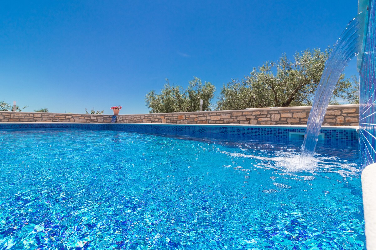 A clear swimming pool is presented with sparkling blue water, reflecting the bright sky. The pool area is bordered by a natural stone wall and features a gentle water feature cascading into the pool, surrounded by greenery and trees in the background.