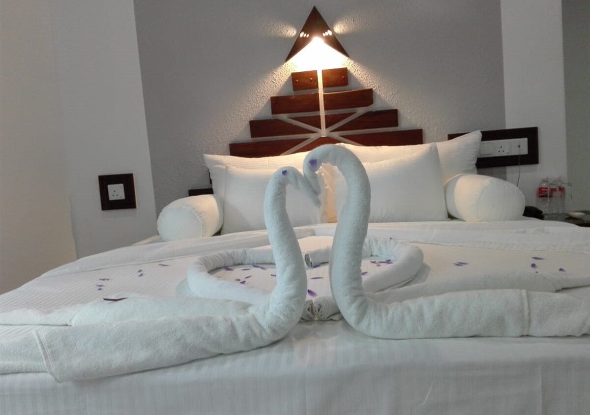 A neatly made bed is centered in the room, adorned with white linens and decorative pillows. Two swan-shaped towel sculptures are created on the bed, accompanied by scattered flower petals, adding a touch of elegance. A soft light fixture is positioned above, enhancing the tranquil ambiance.