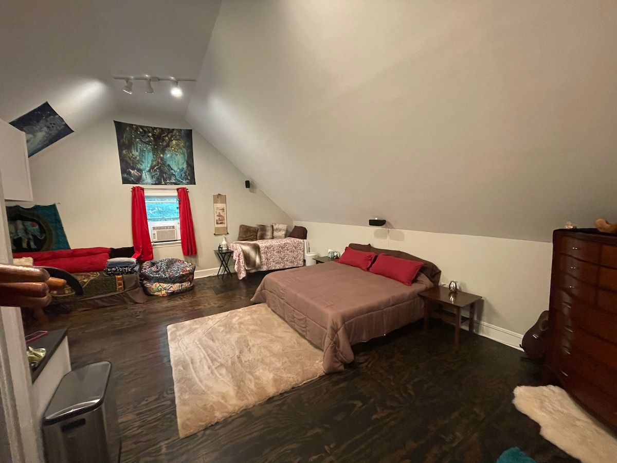 A spacious studio features a sloped ceiling, a bed with decorative pillows, and a cozy area rug. A soft blanket is draped over a second sleeping area, while a colorful seating option and various artworks enhance the decor. Natural light filters through a window, complemented by red curtains.