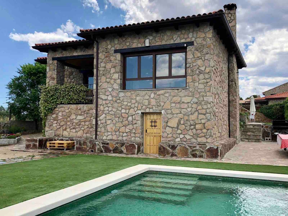 The rustic stone house features large windows offering natural light and views of the surrounding grounds. A welcoming wooden door is visible, leading to a spacious entrance. A well-maintained pool is positioned in the foreground, surrounded by neatly trimmed grass.