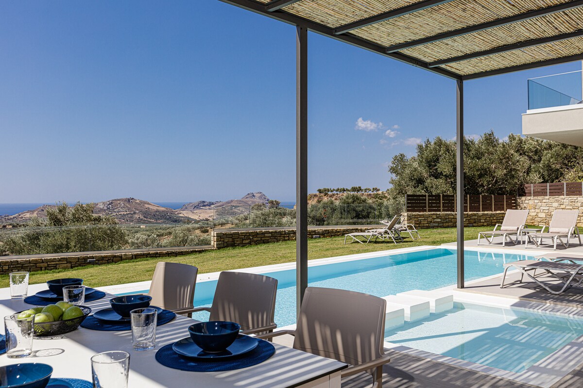 An outdoor dining area is set with tableware, overlooking a private swimming pool and scenic views of the hills and coastline. The shaded terrace features modern furniture, while sun loungers are positioned nearby for relaxation under the clear blue sky.