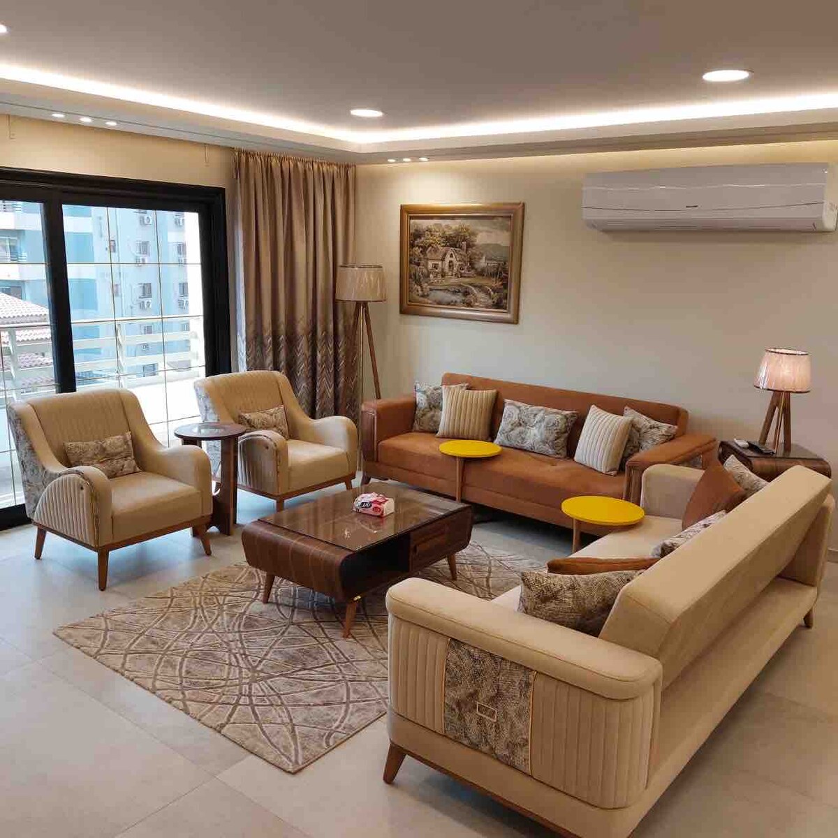 A comfortable living area features a variety of seating options including a spacious sofa and armchairs. Warm lighting from lamps creates a soft ambiance. A coffee table sits on a patterned rug, with large windows providing natural light and views of the surrounding area.