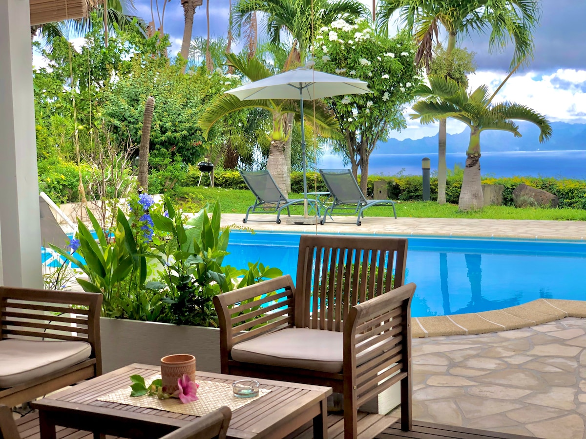 An outdoor seating area features wooden furniture positioned beside a lush garden and a clear pool. A sun umbrella is placed next to lounge chairs, surrounded by tropical plants, enhancing the serene atmosphere and inviting relaxation.