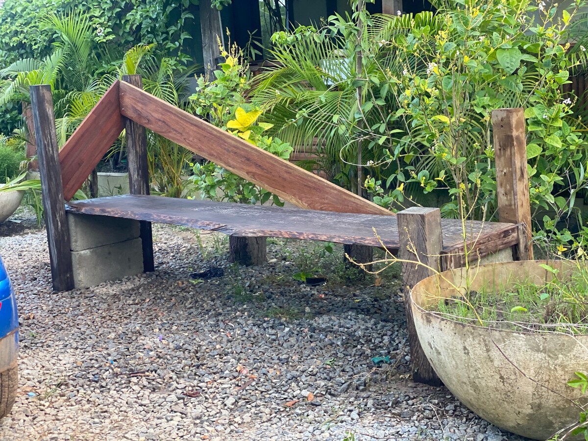 A rustic wooden bench is positioned amidst a natural setting. The bench's slanted design features sturdy wooden supports, while surrounding greenery and tropical plants add a lush backdrop. Gravel covers the ground beneath, contributing to the outdoor ambiance.