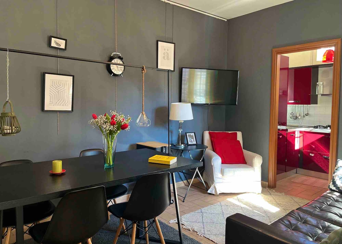 An inviting living space features a large dining table surrounded by black chairs, with a vibrant bouquet of flowers in the center. A comfortable white armchair and a stylish lamp are positioned nearby, while a TV mounted on the wall offers entertainment. The kitchen is visible in the background, showcasing red cabinetry.