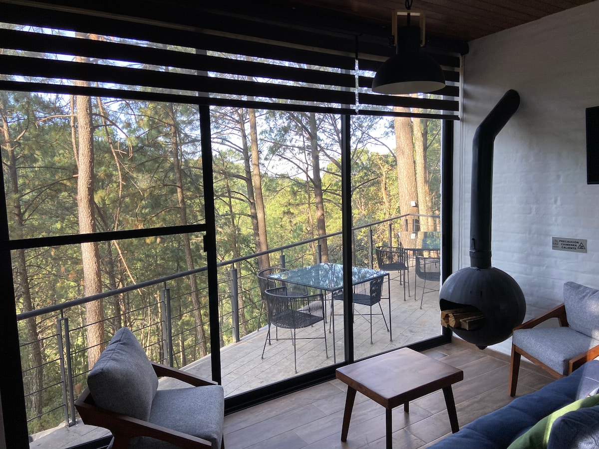 Floor-to-ceiling windows offer views of the peaceful forest, while a cozy seating area features two armchairs and a wooden coffee table. An elegant wood-burning stove adds warmth, and a wooden deck outdoors is furnished with a dining set, perfect for enjoying nature.