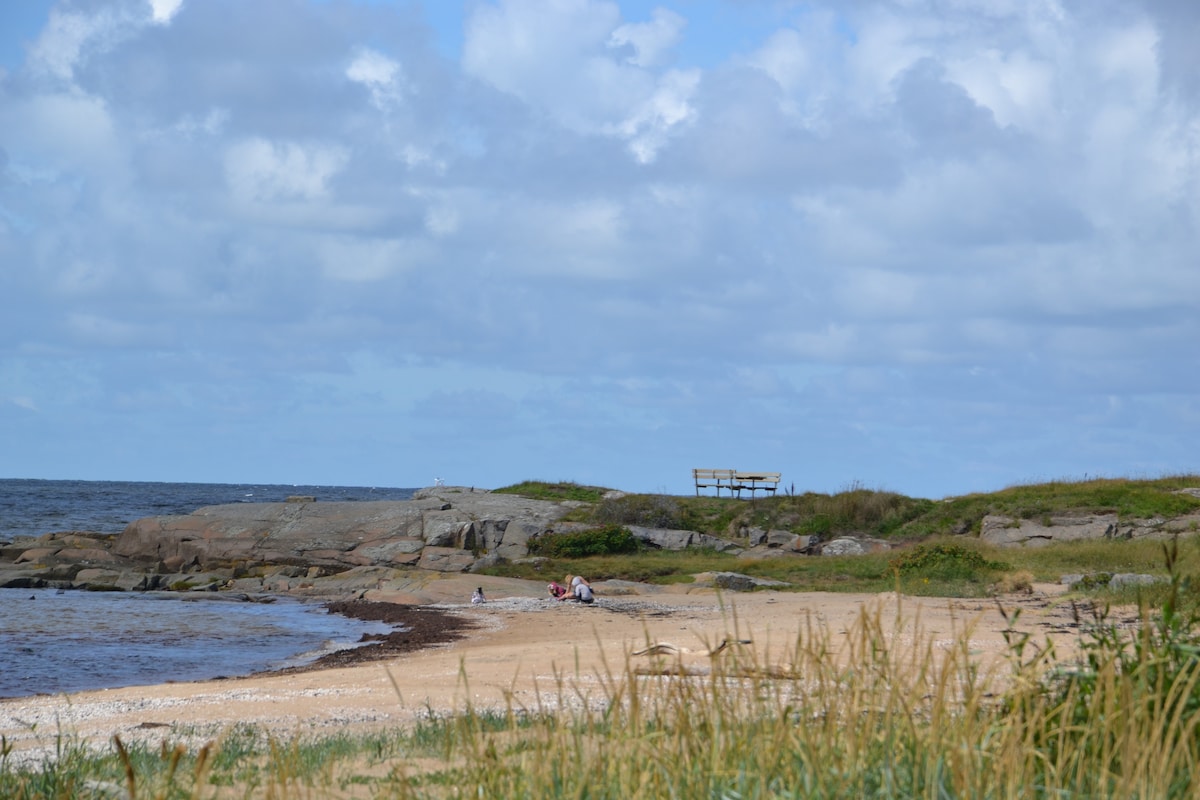 The sandy beach is bordered by smooth rocks and patches of grass, with gentle waves lapping at the shore. A few people are seen enjoying the beach, and a wooden structure is positioned in the background, facing the water under a partly cloudy sky.