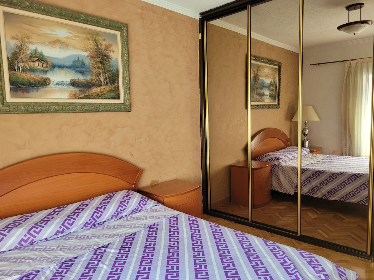 A cozy bedroom features a spacious bed covered with a patterned purple and white blanket. A mirrored wardrobe reflects the room's warm tones and includes a bedside table and lamp. Art depicting a serene landscape adorns the wall, contributing to the room's inviting ambiance.