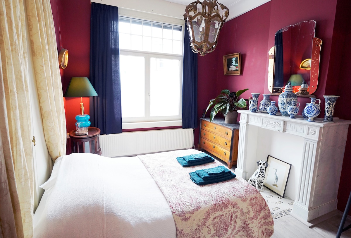 The bedroom features rich burgundy walls and a spacious bed with a decorative coverlet. A traditional candle chandelier hangs overhead, and a large window dressed with blue curtains allows natural light to filter in. An antique dresser and a fireplace with decorative ceramics add charm to the space.