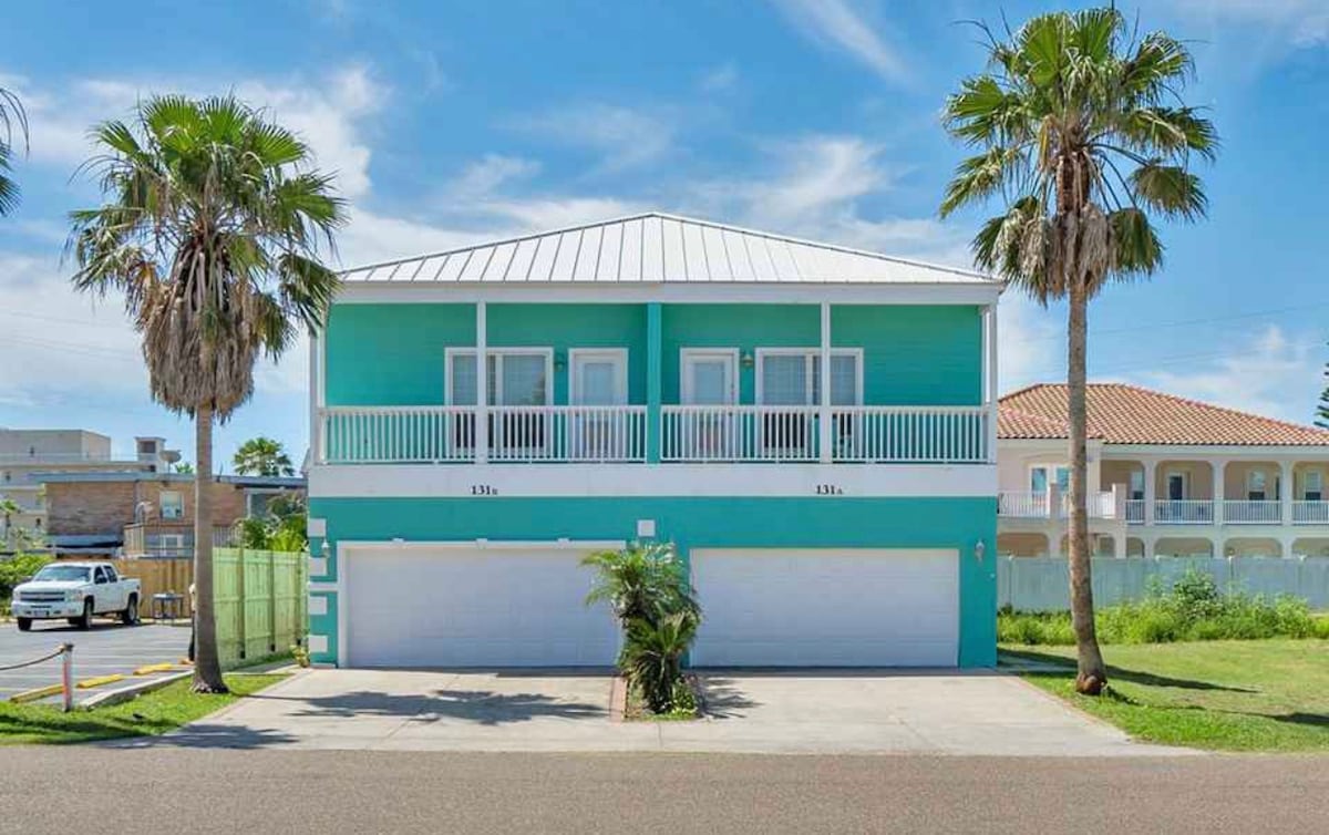 A two-story turquoise house is framed by tropical palm trees, with a spacious driveway leading to a two-car garage beneath a covered entrance. Large windows offer a view of outdoor spaces, and the well-maintained surrounding area contributes to the inviting curb appeal.