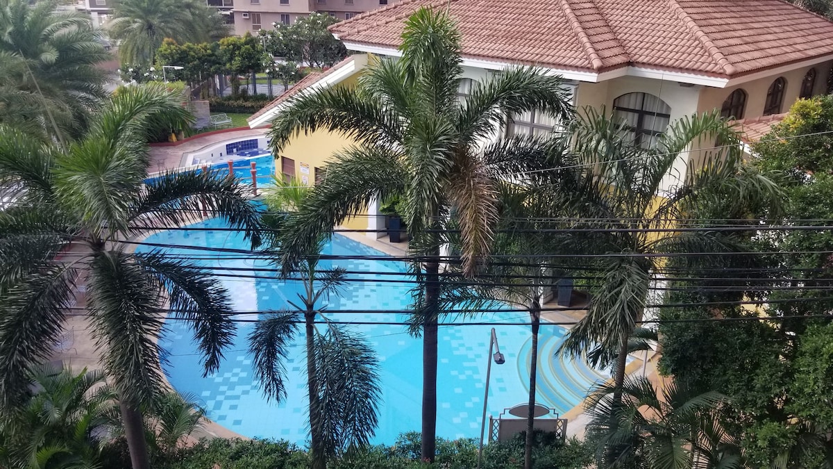 A view of a well-maintained pool area surrounded by lush palm trees. The water reflects the sunlight, and colorful tiles enhance the inviting appearance of the pool. A building with a reddish-tiled roof is visible in the background, complementing the serene environment.