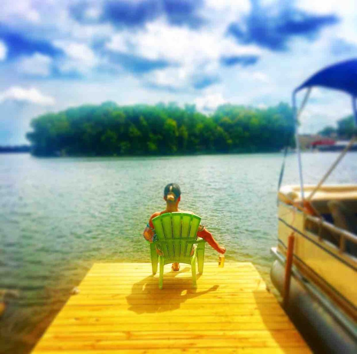 A person sits in a green Adirondack chair on a wooden dock, facing the calm waters of Lake Shafer. Lush greenery can be seen across the lake, with a boat anchored nearby. The sky is partly cloudy, adding to the serene lakeside atmosphere.
