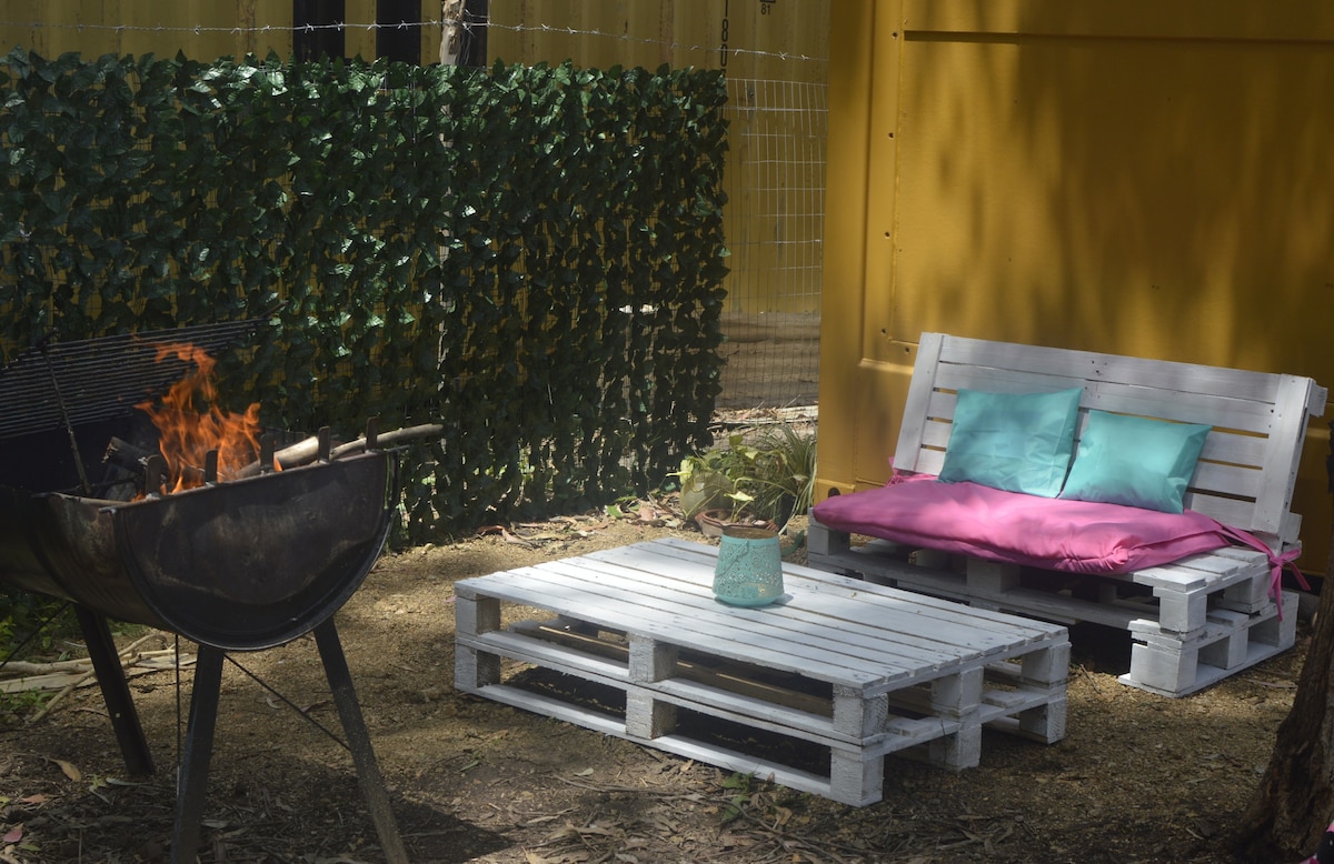 An outdoor seating area features a pallet sofa with teal cushions and a pink throw, positioned next to a small, simple coffee table. A fire pit with flames is visible, surrounded by greenery that provides a natural backdrop.